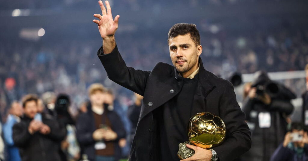 Spain footballer Rodri looking to his right and raising his right hand in a football stadium while wearing a black jacket and shirt and holding the golden Ballon d'Or trophy with teammates blurrily visible behind him