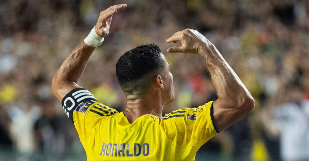 Il calciatore portoghese Cristiano Ronaldo alza entrambe le braccia dando le spalle alla telecamera in uno stadio mentre gioca per la squadra della Saudi Pro League Al Nassr Football Club con indosso una maglia gialla e blu