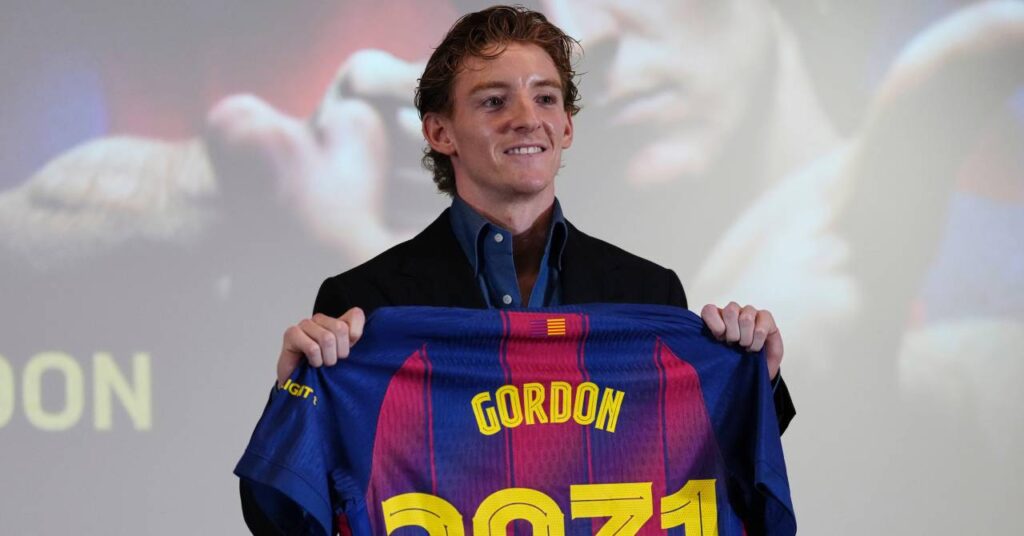 England footballer Anthony Gordon smiling and holding up a red and blue striped shirt with his surname and the number 2031 on the back in yellow while being presented as a player for La Liga team Barcelona Football Club and wearing a black suit jacket and dark blue shirt