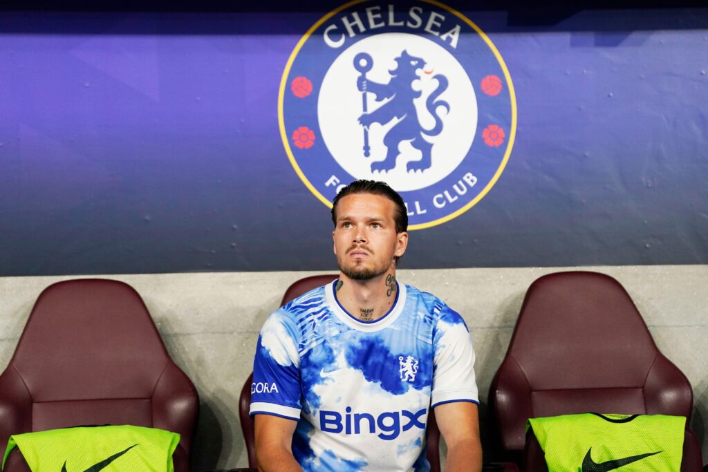 Ukraine footballer Mykhailo Mudryk sitting on a bench in a technical area looking sternly upwards with the circular blue, white and yellow badge of Premier League team Chelsea Football Club behind him