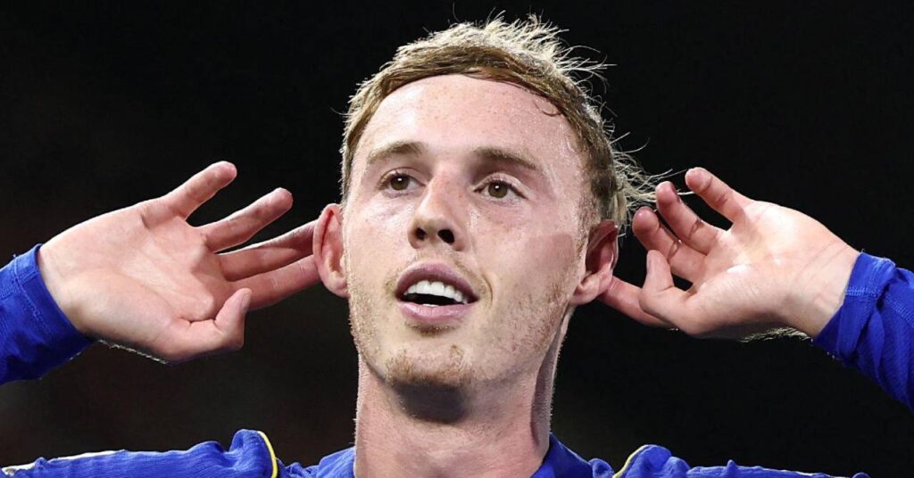England footballer Cole Palmer holding his hands behind his ears while celebrating a goal under floodlights in a stadium while playing for Premier League team Chelsea Football Club and wearing a blue shirt