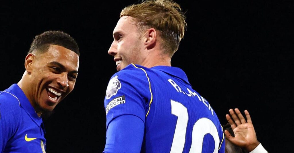 England footballer Cole Palmer looking to his left while smiling and being congratulated by teammate Morgan Rogers under floodlights in a stadium while playing for Premier League team Chelsea Football Club and wearing a blue shirt with his name and number 10 in white on the back