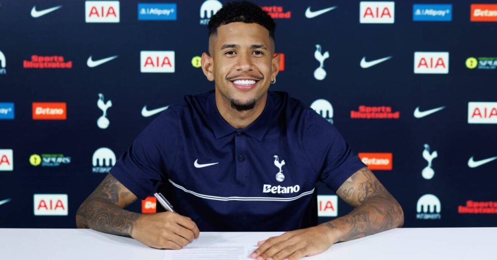 Brazil footballer Savinho posing at a white table to sign a contract at Premier League team Tottenham Hotspur Football Club while wearing a dark blue Spurs shirt in front of club and sponsor logos