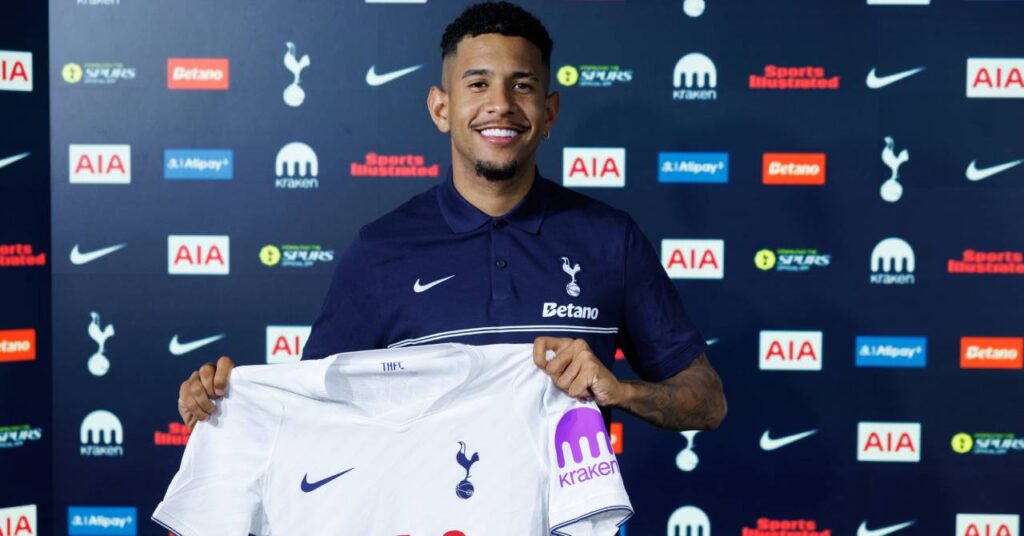 Brazil footballer Savinho posing with a white shirt to sign a contract at Premier League team Tottenham Hotspur Football Club while wearing a dark blue Spurs shirt in front of club and sponsor logos