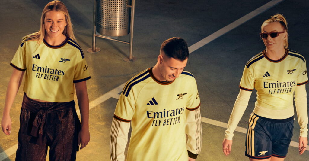 Footballers Chloe Kelly, Alessia Russo and Gabriel Martinelli standing in a shadowy room in the third kit of Premier League team Arsenal Football Club which is coloured yellow with blue trim