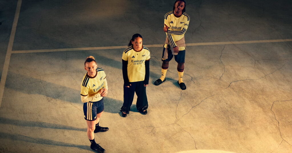Footballers Chloe Kelly, Olivia Smith and Eberechi Eze standing in a shadowy room in the Arsenal Football Club third kit of yellow with blue trim