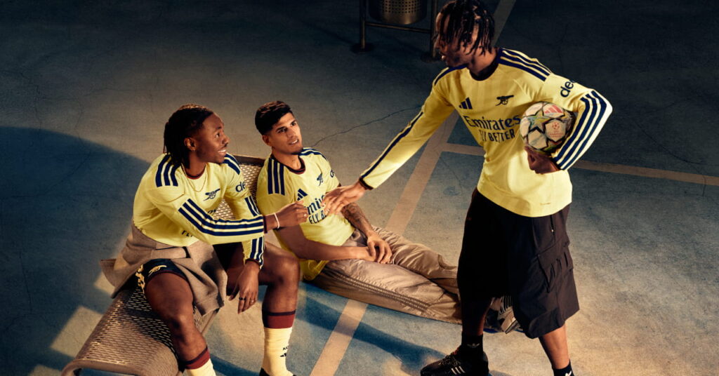 Footballers Noni Madueke, Piero Hincapie and Eberechi Eze standing in a shadowy room in the third kit of Premier League team Arsenal Football Club which is coloured yellow with blue trim