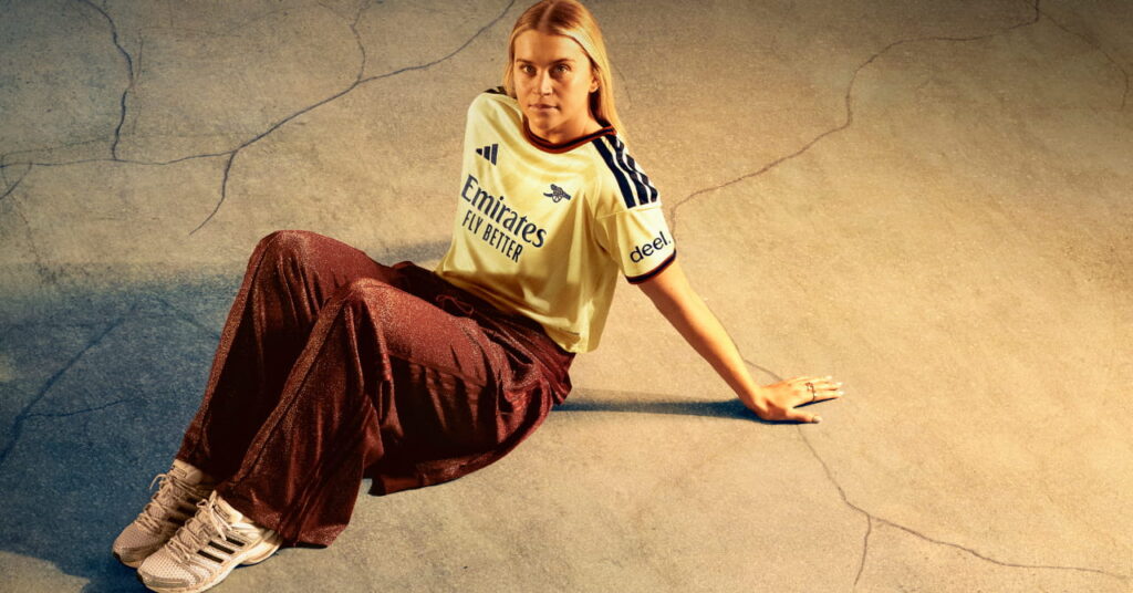 Footballer Alessia Russo sitting in a shadowy room in the third kit of Premier League team Arsenal Football Club which is coloured yellow with blue trim