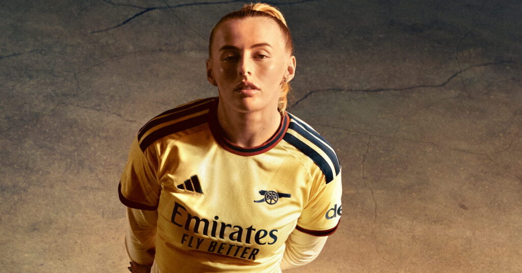 England footballer Chloe Kelly standing in a shadowy room in the third kit of Premier League team Arsenal Football Club which is coloured yellow with blue trim