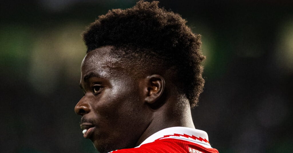 O jogador de futebol Bukayo Saga é mostrado com seu nome e número em branco, de costas para a câmera, no Arsenal Football Club, predominantemente vermelho e branco, enquanto joga pelo time da Premier League no estádio à noite.