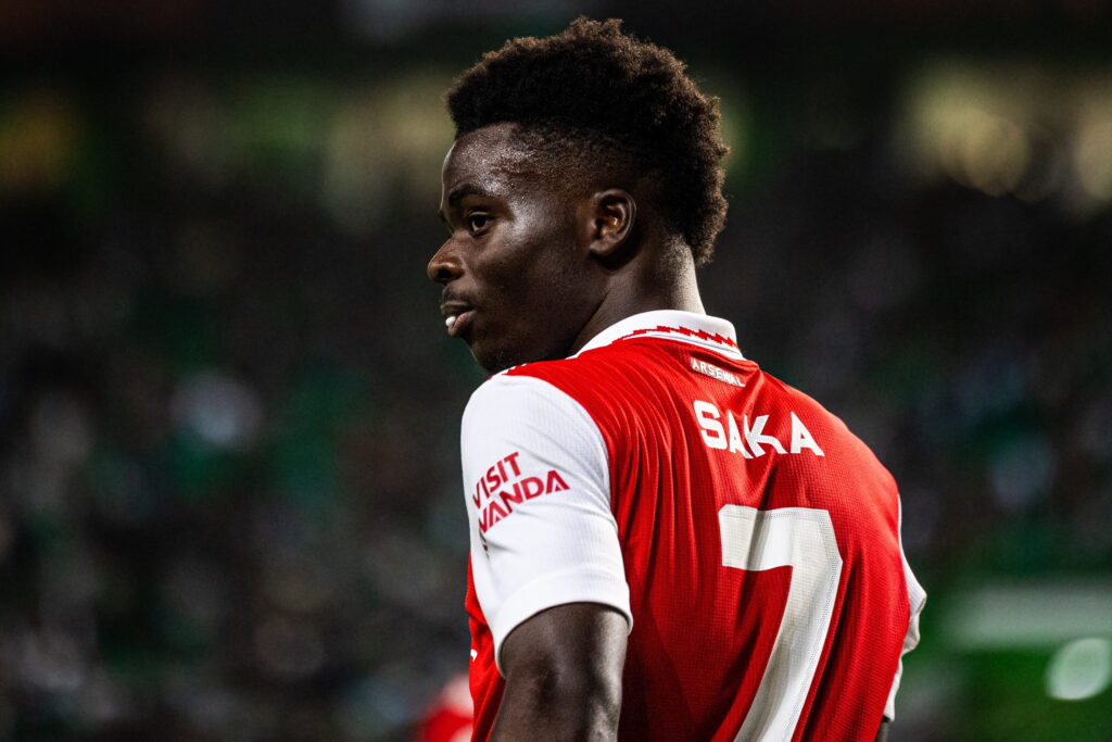 Footballer Bukayo Saka is shown with his back to the camera and his name and number in white on a predominantly red and white Arsenal Football Club while playing for the Premier League side in a stadium at night