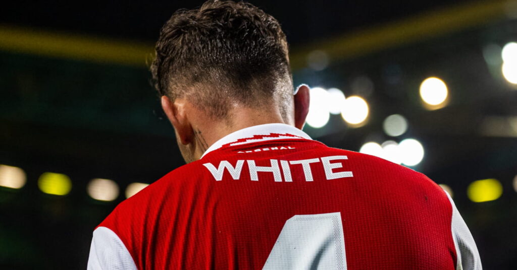 Footballer Ben White is shown with his back to the camera and his name and number in white on a predominantly red and white Arsenal Football Club while playing for the Premier League side in a stadium at night