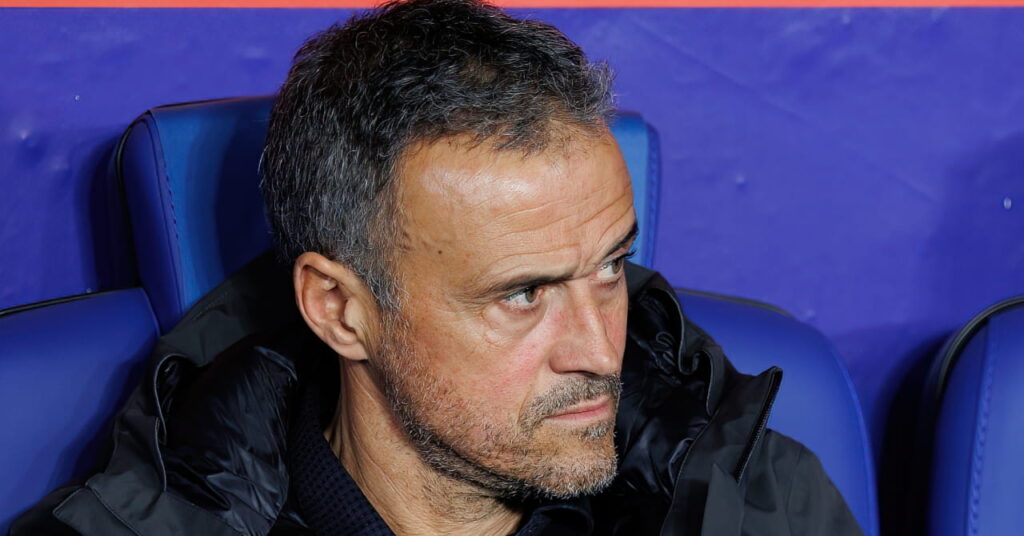 O técnico de futebol Luis Enrique está sentado em um assento azul em uma área técnica e olha para a esquerda, vestindo um casaco escuro como técnico do Paris Saint-Germain ou do clube de futebol PSG.