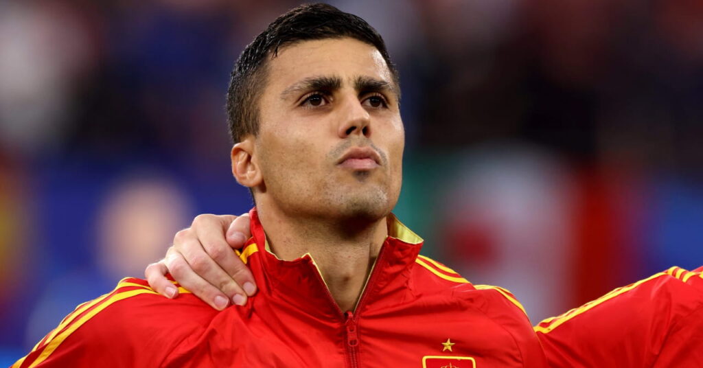 Spain footballer Rodri standing and linking arms with teammates for the national anthem in a football stadium in a red jacket with yellow trim