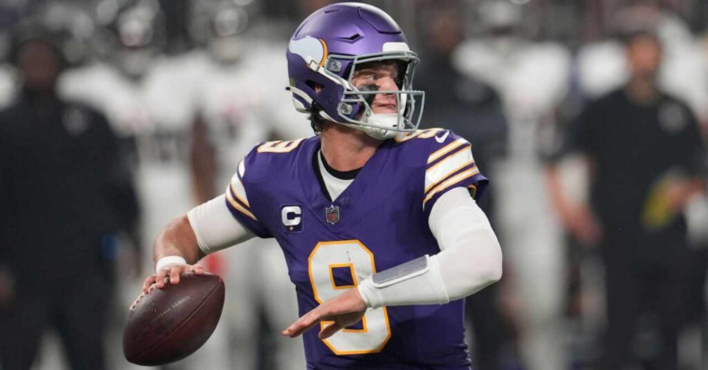 O quarterback de futebol americano da National Football League, J.J. McCarthy, vestindo uma camisa roxa e capacete do Minnesota Vikings com detalhes amarelos, brancos e pretos, e o número nove na frente com uma bola de futebol americana marrom na mão esquerda, olhando para a esquerda no estádio.
