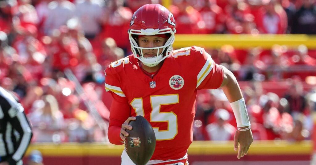 O jogador de futebol americano Patrick Mahomes segura uma bola marrom da National Football League enquanto o quarterback joga com um uniforme vermelho e branco em um campo pelo time Kansas City Chiefs NFL.