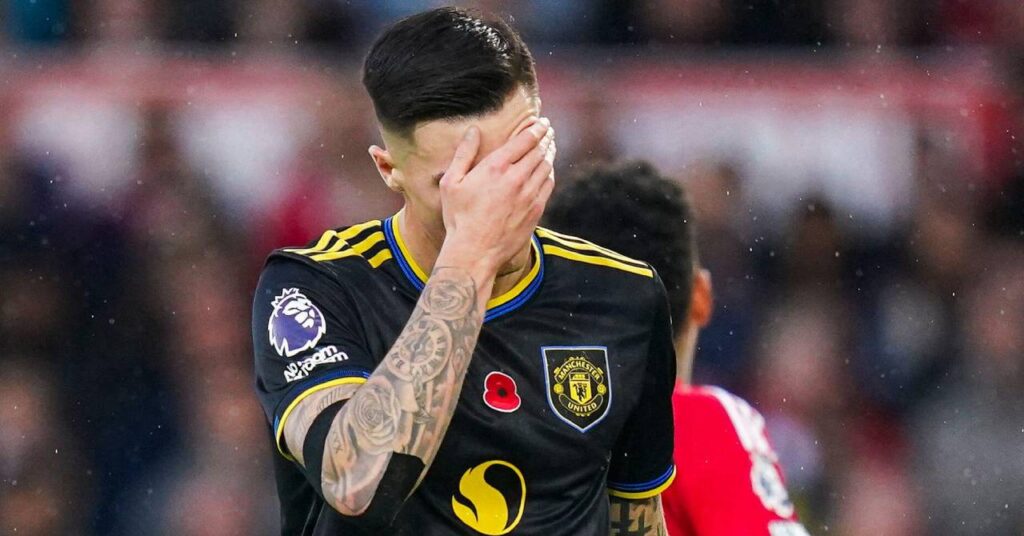 Slovenia footballer Benjamin Sesko holding his left arm to his face while playing for Premier League team Manchester United Football Club in a black kit with yellow trim in a stadium with opposition players in red and a crowd blurrily visible behind the forward