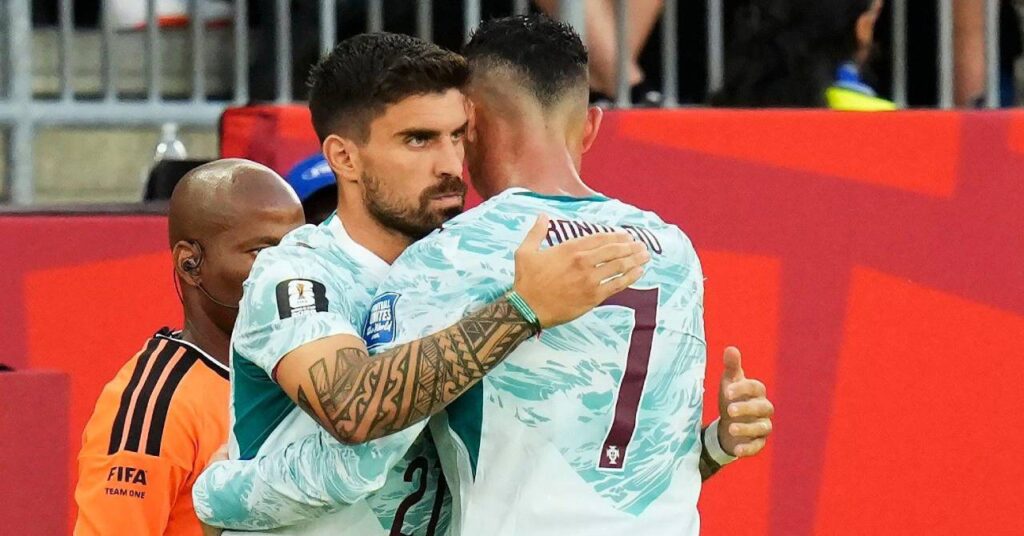 Portugal footballers Cristiano Ronaldo and Ruben Neves embracing on a football pitch while wearing a light green and white kit with their names and numbers in purple on the back in front of a red and metal stand with an official in an orange and black kit nearby
