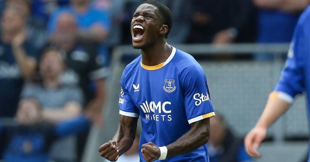O jogador de futebol Thiarno Barry, com uma camisa azul com detalhes em branco e amarelo e o emblema do clube e o logotipo Castor no peito, cerra os punhos em comemoração ao comemorar um gol pelo Everton Football Club, da Premier League.