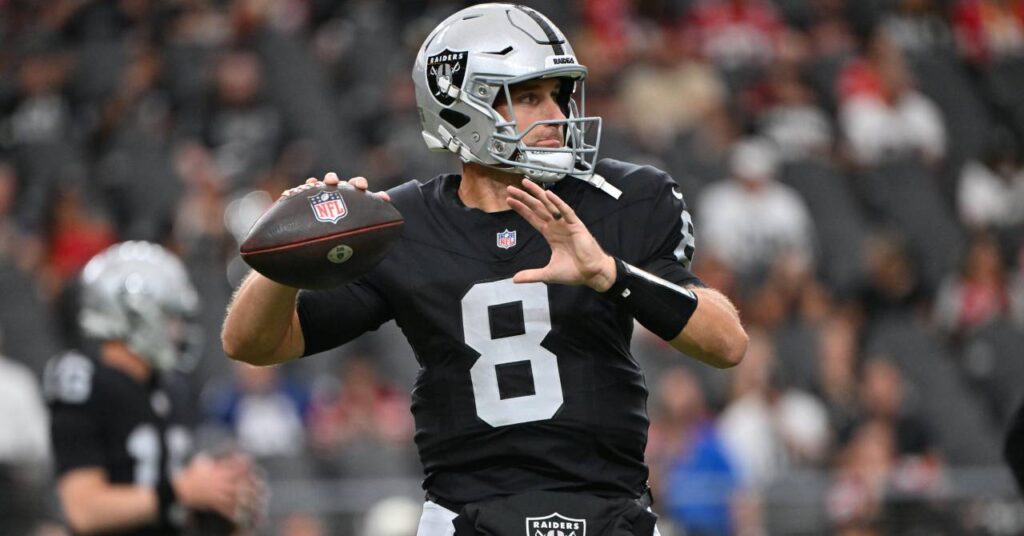 American footballer Kirk Cousins playing for National Football League team the Las Vegas Raiders in the NFL and raising his left hand to throw a ball while wearing a silver helmet and black shirt with his number eight in white on the shirt in a stadium with a crowd blurrily visible in the background