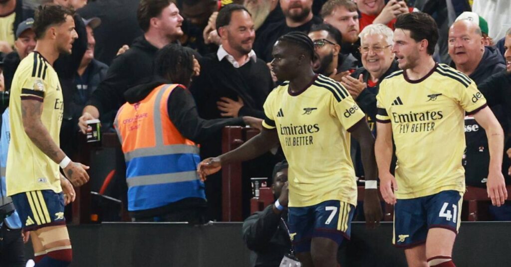 England footballers Declan Rice and Bukayo Saka looking to their right with Ben White looking across at them on a pitch in a yellow shirt with blue trim, blue shorts and red socks while playing for Premier League team Arsenal Football Club in front of a celebrating crowd