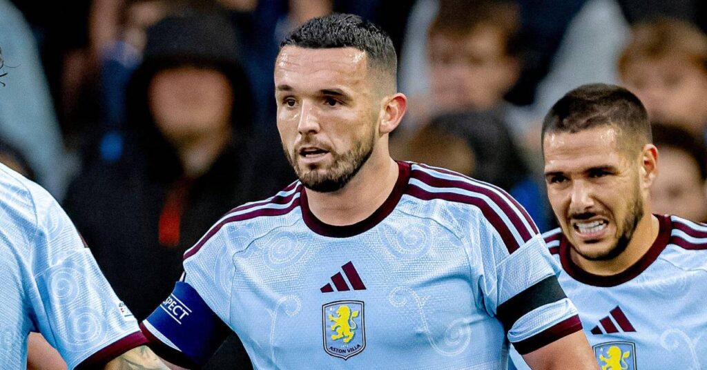 O jogador de futebol escocês John McGinn, vestindo uma camisa azul clara, joga pelo Aston Villa Football Club, da Premier League, com o companheiro de equipe Emiliano Buendia e uma leve multidão atrás dele.