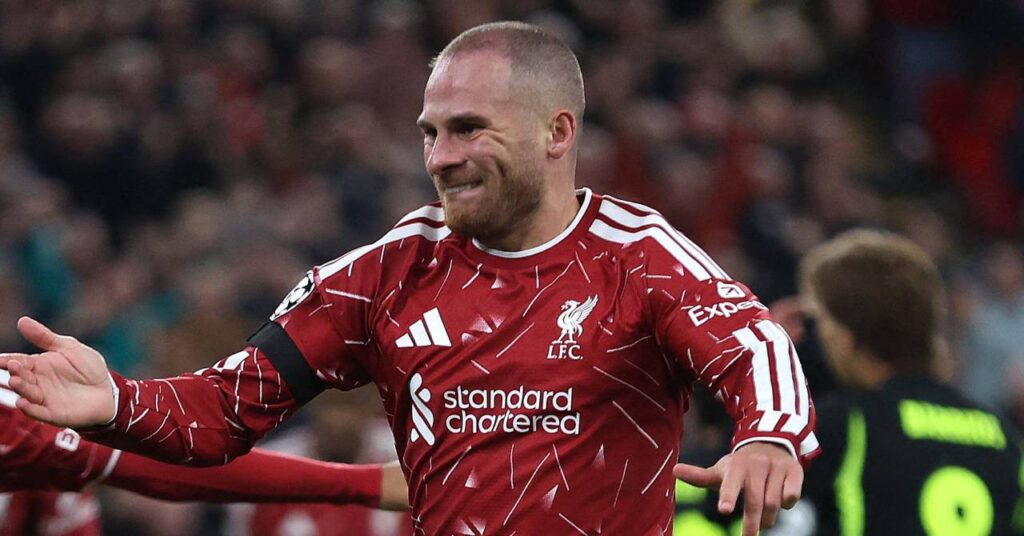 Argentina footballer Alexis Mac Allister raising his right hand to celebrate a goal while playing for Premier League team Liverpool Football Club in a red shirt with white trim and a Champions League badge on the right arm with a crowd blurrily visible in the background