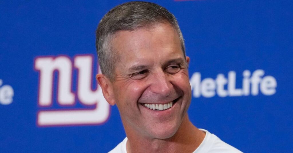 O técnico de futebol americano John Harbaugh, sorrindo, olha para a esquerda e fala em uma entrevista coletiva com o time da National Football League, New York Giants, vestindo uma blusa branca na frente de uma placa azul com as palavras NY e MetLife em letras brancas e vermelhas.