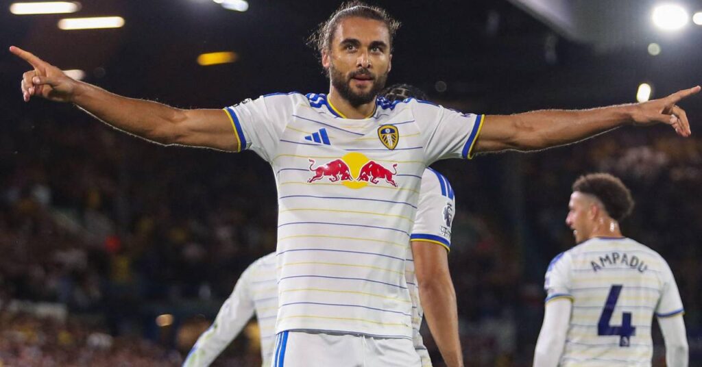 Footballer Dominic Calvert-Lewin speading his arms in a stadium at night to celebrate a goal against Newcastle United while playing for Premier League team Leeds in a white kit with blue trim with a crowd visible