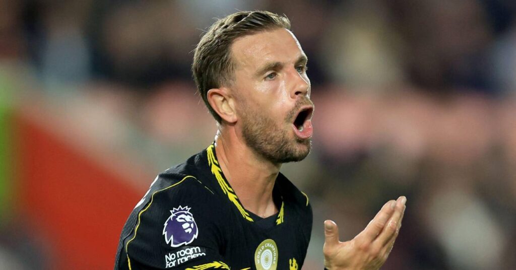 O jogador de futebol inglês Jordan Henderson gesticula com a mão direita enquanto joga pelo Chelsea Football Club, clube da Premier League, vestindo uma camisa preta com detalhes amarelos e um símbolo circular no peito representando o clube campeão da Copa do Mundo