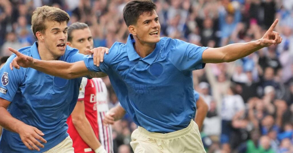Footballer Chema Andres raising both arms and his index fingers to celebrate a goal while playing for Premier League club Brighton and Hove Albion Football Club in a blue shirt and white shorts in front of a blurrily visible crowd with Croatia footballer Luka Vuskovic congratulating him