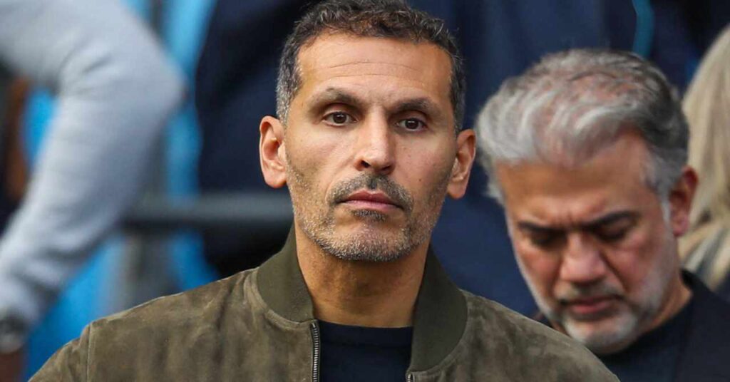 Premier League team Manchester City Football Club chairman Khaldoon Al Mubarak looking forward while standing at the Etihad Stadium wearing a green jacket and black shirt with other members of a crowd visible behind the chief executive officer and managing director of Mubadala Investment Company