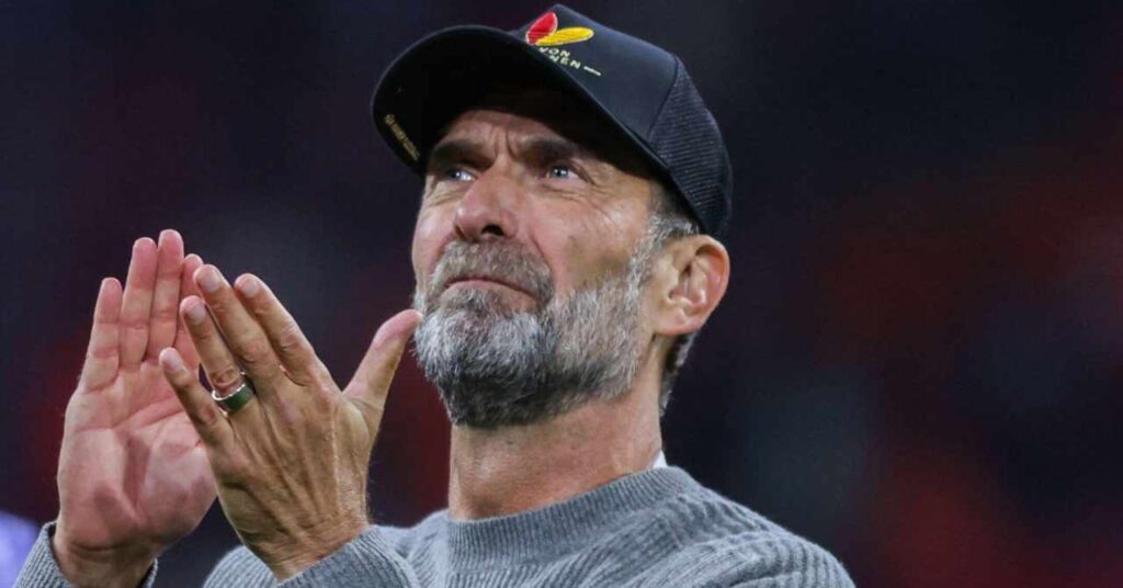 O técnico de futebol Jurgen Klopp, usando um boné de beisebol preto da Adidas com o logotipo da empresa em preto, vermelho e amarelo, olha para a direita em um campo em seu papel de técnico e aplaude.