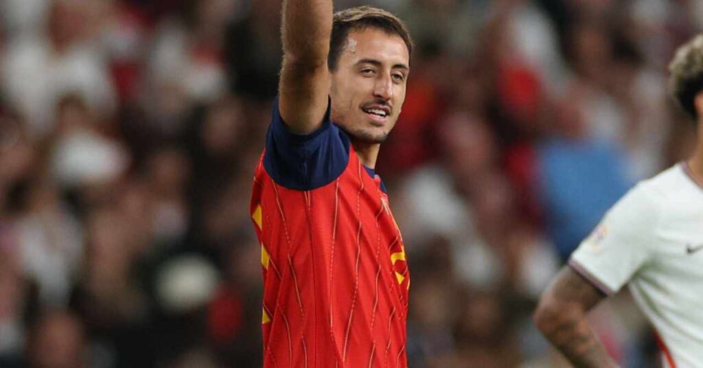 O jogador de futebol da La Liga da Real Sociedad, Mikel Oyerzabal, comemora um gol com o braço direito levantado enquanto joga pela seleção espanhola de futebol com uma camisa vermelha com detalhes azuis e listras amarelas.