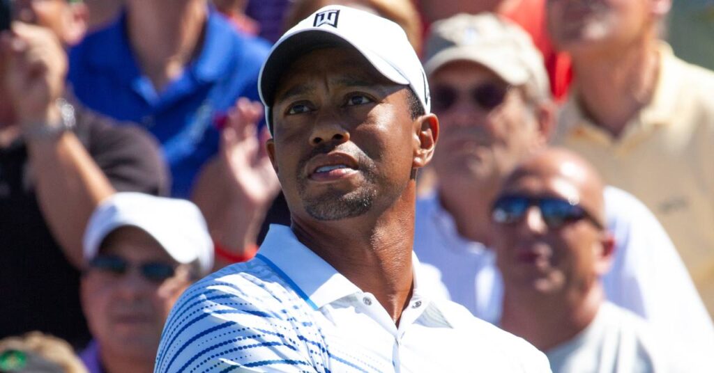 Golf player Tiger Woods taking a shot in front of a crowd while wearing a white baseball cap and polo shirt
