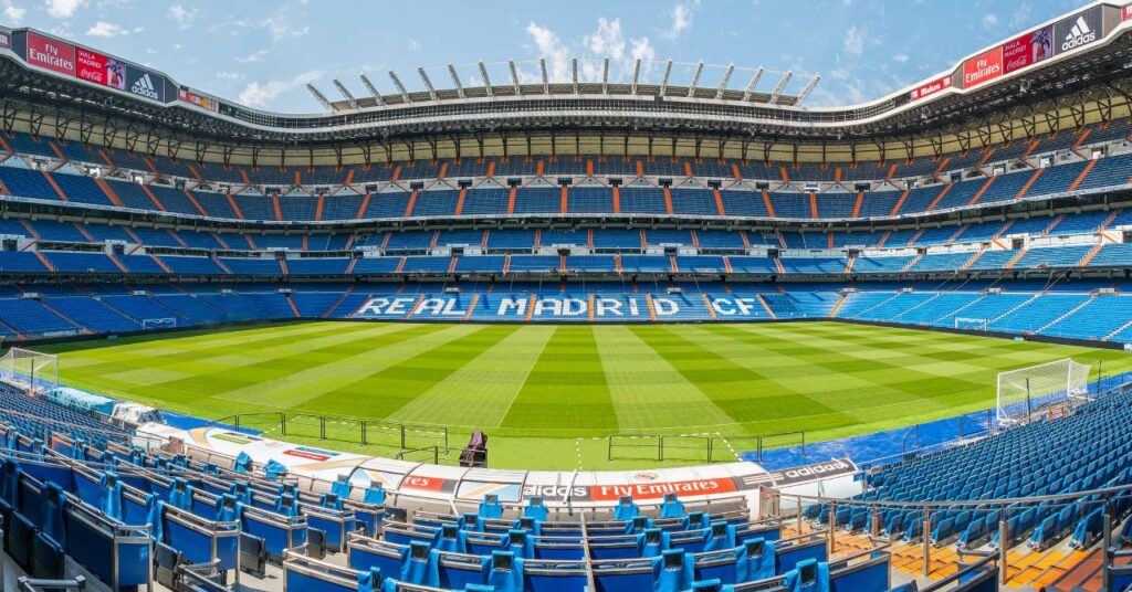 The Santiago Bernabeu Stadium home of La Liga team Real Madrid Football Club showing three tiers of blue seats around a green pitch with the club name spelled out in white in the seats on one side of the lower tier
