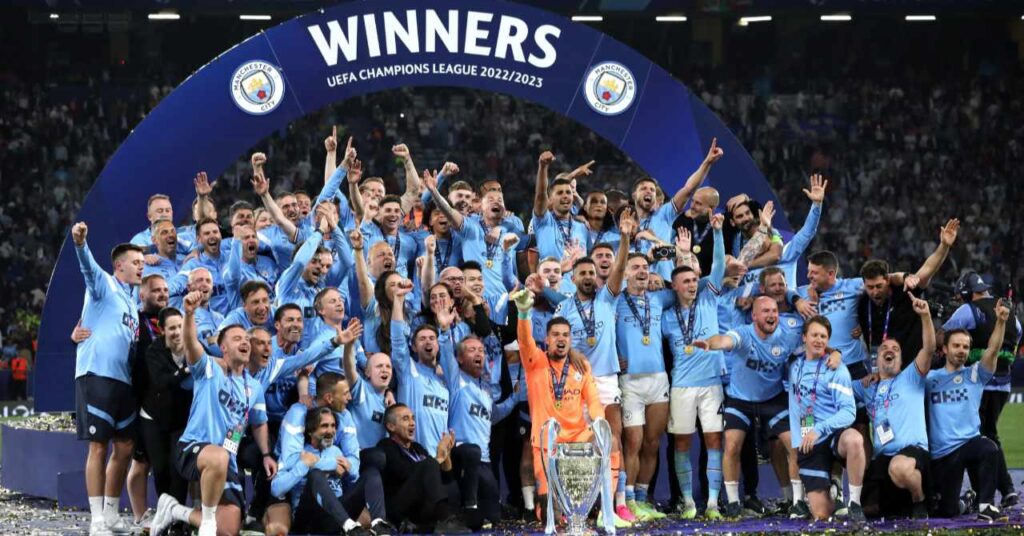 Manchester City Football Club players posing with the UEFA Champions League trophy under a banner saying winners UEFA Champions League 2022/23 while wearing Man City light blue shirts and white shorts in a stadium
