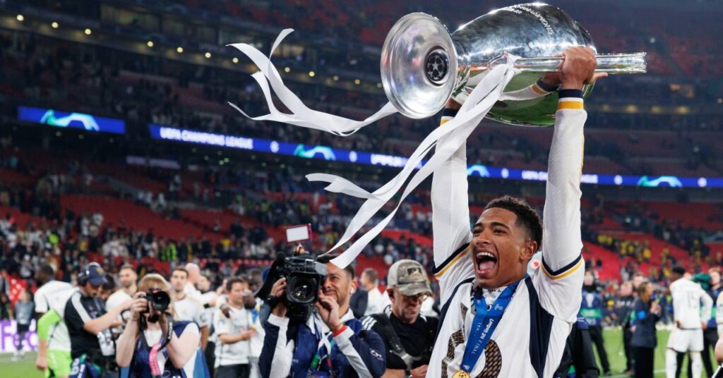 England footballer Jude Bellingham holding the silver Champions League trophy horizontally in a stadium while playing for La Liga team Real Madrid Football Club in a white shirt with yellow and dark blue trim