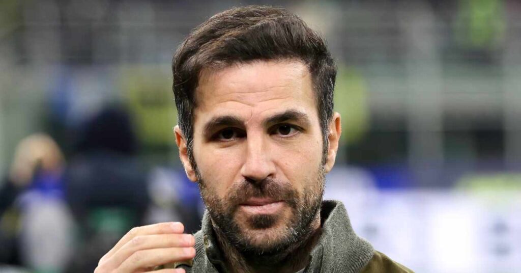 Football manager Cesc Fabregas raising his right hand to his face in a stadium while wearing a grey and olive jacket in his role as head coach of Serie A football team Como Football Club.