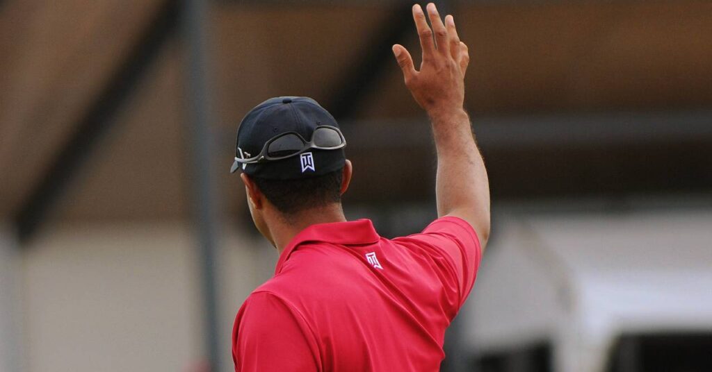 Golf player Tiger Woods using his right hand to wave to a crowd with his back to the camera while wearing a black baseball cap and red polo shirt