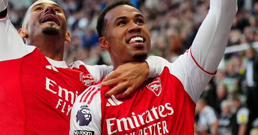 Brazilian footballer Gabriel Magalhaes looking up and smiling and raising his left arm to celebrate a goal for Premier League team Arsenal Football Club as teammate and French footballer William Saliba hugs him around the neck from his right. They wear a red home shirt with white sleeves and the Premier League Lion logo on the sleeves and the words Emirates Fly Better and the club and Adidas logos in white on the chest with a crowd visible in the background.