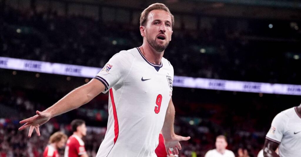 Footballer Harry Kane celebrating a goal by spreading his arms while running to his left while the player for Bundesliga team Bayern Munich Football Club is playing in the England national football team home shirt of white with dark blue details with the Nike logo and Three Lions crest on the chest above his number nine in red with teammates, opposition players in red and a crowd blurrily visible behind him.