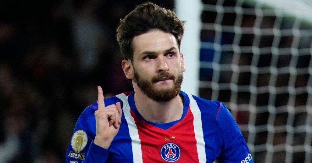 Georgia's footballer Khvicha Kvaratskhelia raises his right hand and index finger in celebration of a goal while playing for Ligue 1 club Paris Saint Germain Football Club while wearing a blue, red and white PSG shirt at a stadium in front of a goal post and a white goal net. The Ligue 1 logo is visible on the right arm and the club badge is on the chest with a graphic of the Eiffel Tower in France.
