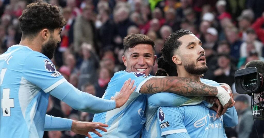 O jogador de futebol francês Ryan Sergi, à direita, comemora um gol do Manchester City Football Club, da Premier League, usando uma guarnição branca e o logotipo da Etihad Airways no peito e o logotipo do leão da Premier League no braço direito. O companheiro de equipe do Man City e jogador de futebol argentino, Enzo Fernandez, abraça e sorri atrás de Sergi enquanto o jogador de futebol croata Josco Guardiola levanta os dois braços na frente de uma multidão pouco visível.