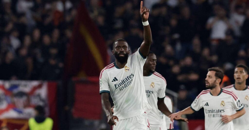 Germany footballer Antonio Rudiger raising his left arm and index finger to celebrate a goal while playing for La Liga team Real Madrid Football Club in a white kit with pink strips on the arms and his number 22 in dark green on the shorts. The words Emirates Fly Better are visible on his chest in dark green under the Adidas logo and club badge.