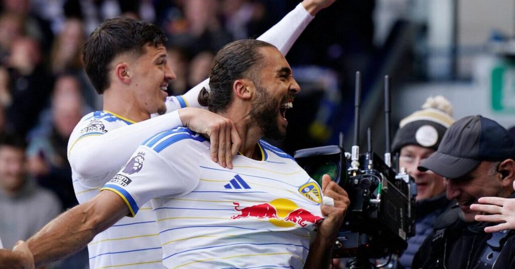 O jogador de futebol Dominic Calvert-Lewin parece deixado em campo, sorri e grita em comemoração a um gol enquanto joga pelo Leeds United, time da Premier League, com uma camisa branca com detalhes azuis e listras amarelas. Calvert Levine é flanqueado por dois operadores de câmera, flanqueados por um companheiro de equipe usando um logotipo vermelho e amarelo da Red Bull, um logotipo azul da Adidas e um emblema do clube no peito.
