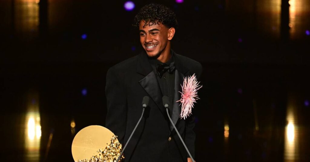 Spain footballer Lamine Yamal smiling to his right on a stage. The player for La Liga team Barcelona is wearing a black suit with a circular gold award in front of him near two microphones and a pink floral decoration to his left in front of dim lighting.